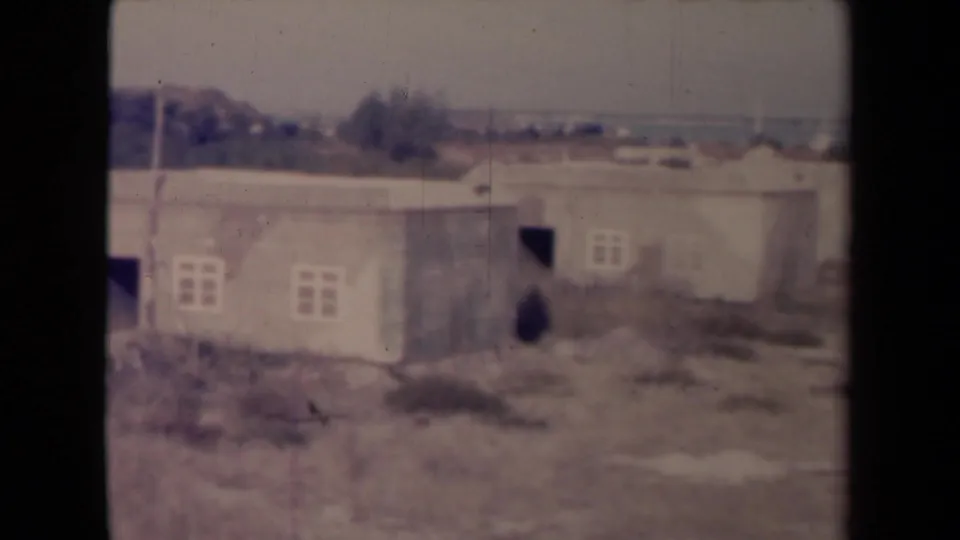 1945 France 8mm view panning along connected low buildings with pale walls, small windows, open doorways, scrub, and distant trees.