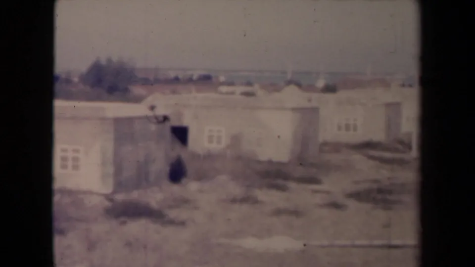 1945 France frame at the end of the pan, showing low rural buildings, an open doorway, small windows, dry grass, and a faint horizon.