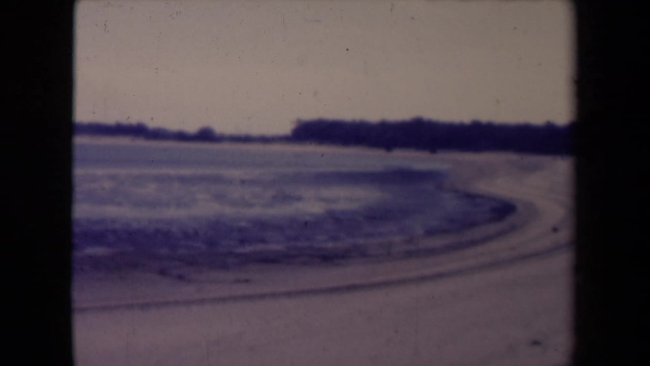 1945 France home-movie frame showing a quiet shoreline, pale water and a dark distant tree line.