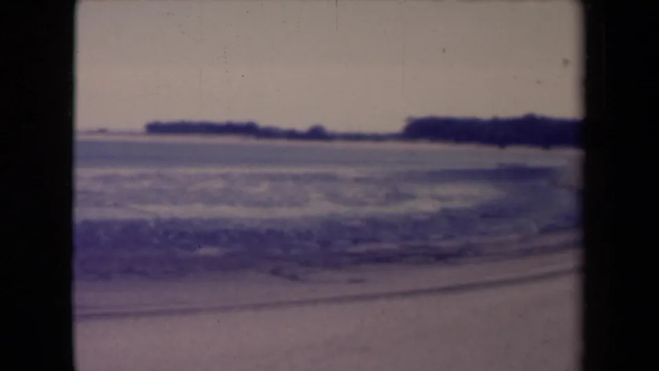 France 1945 8mm frame showing small ocean waves, a sandy beach, and a low distant shoreline under a hazy sky.
