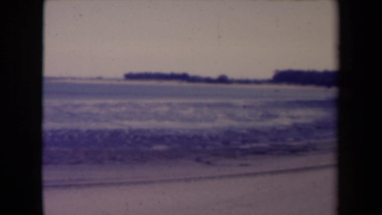 France 1945 8mm frame of choppy coastal water, foreground sand, and dark low land across the horizon.