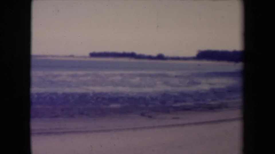 France 1945 8mm frame of choppy coastal water, foreground sand, and dark low land across the horizon.