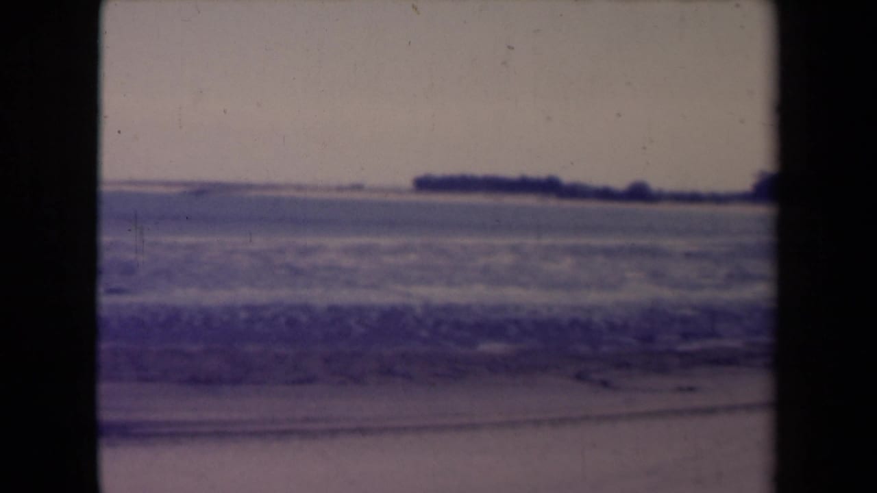 France 1945 8mm frame showing surf near the beach and a dark, low shoreline in the distance.