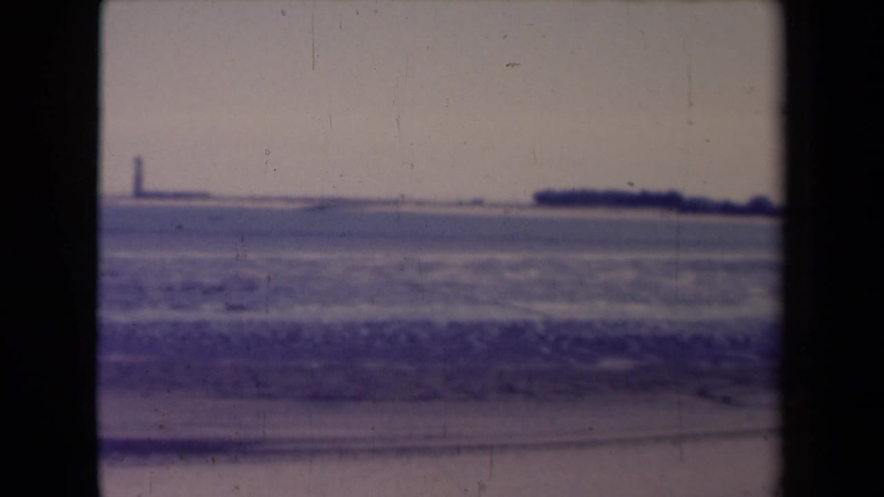 France 1945 8mm frame with beach surf, distant shoreline, and a faint lighthouse-like tower across the water.