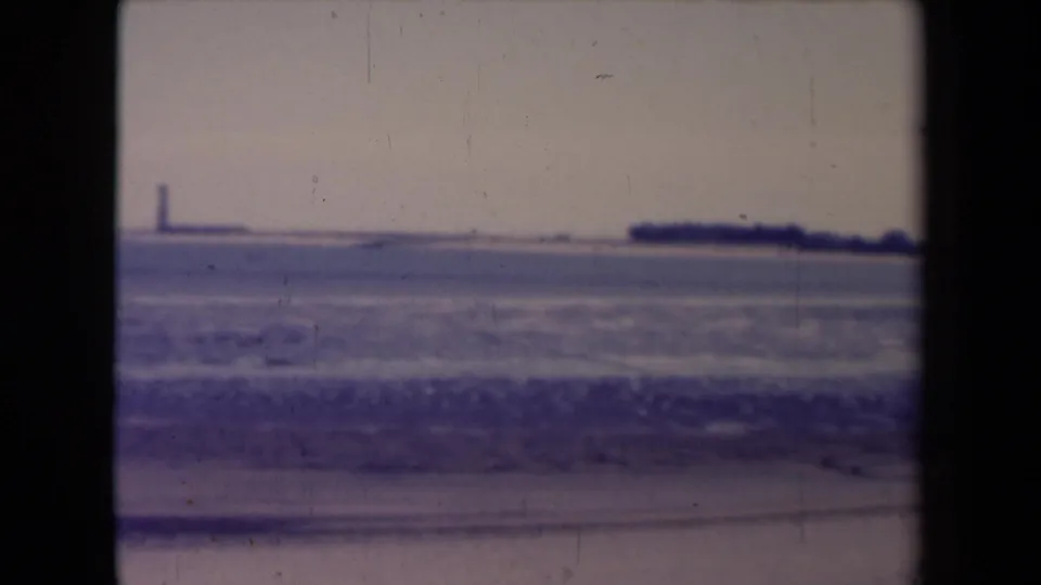 France 1945 8mm frame with beach surf, distant shoreline, and a faint lighthouse-like tower across the water.