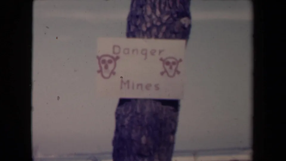 Centered 8mm view of the hand-drawn Danger Mines sign on a tree, with skull markings and muted snowy ground beyond.