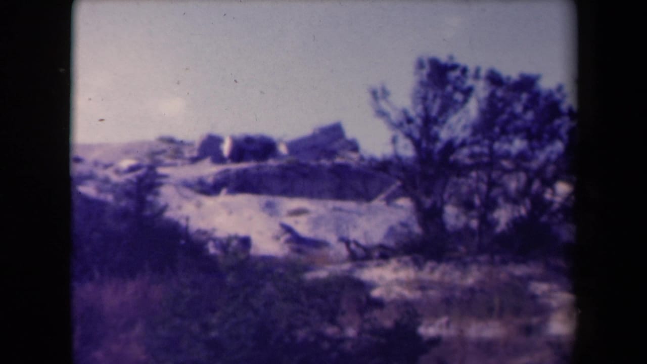 Rocky rise with low structures, scrub, and a tree at the right edge.
