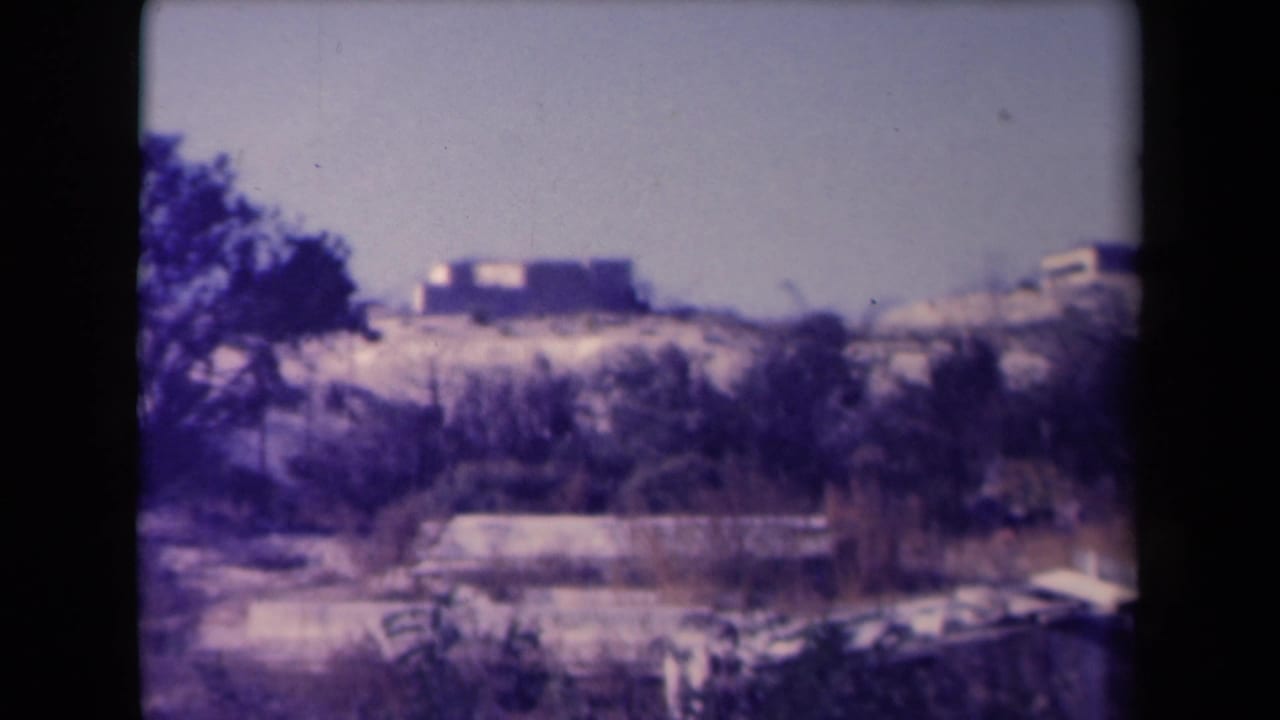 Aged color 8mm frame showing distant low buildings or ruins across scrubby dry ground in France.