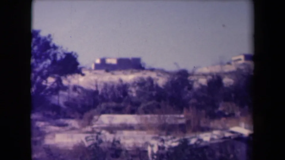 Aged color 8mm frame showing distant low buildings or ruins across scrubby dry ground in France.