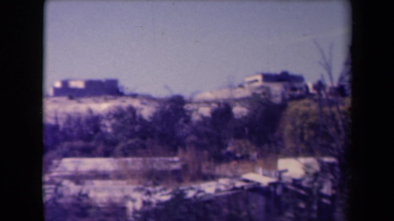 Sandy ridge with distant homes, brush, foreground clutter, and pale sky.