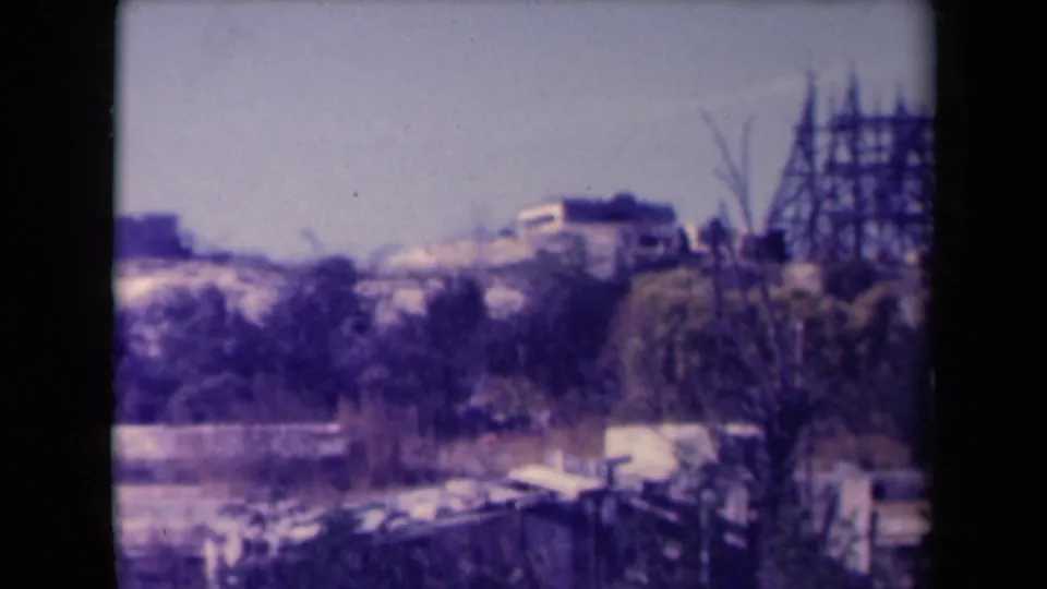 1945 France 8mm frame with distant hillside buildings and a tall wooden scaffold appearing on the right.