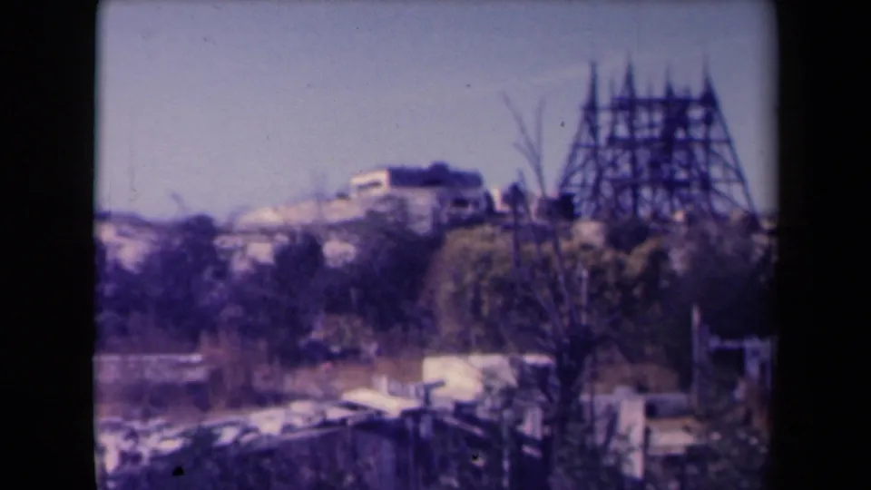 1945 France home-movie frame showing sandy hillside homes and a prominent wooden scaffold beyond foreground brush.