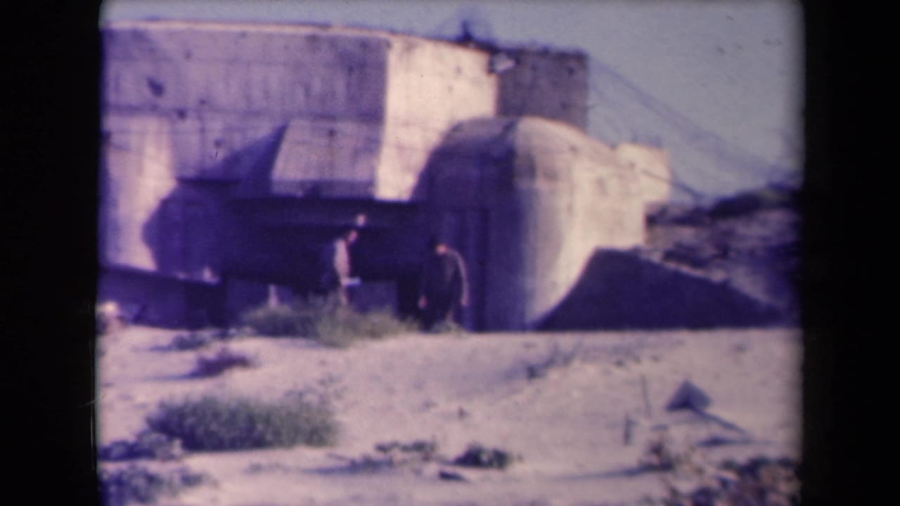 Two small figures stand or walk near a large concrete bunker in bright sun.