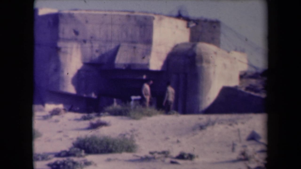 Two figures walk near the rounded side of a concrete bunker structure.