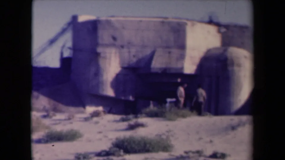 1945 France 8mm frame showing a broad concrete bunker facade with two figures near a shadowed recess and scrubby ground.