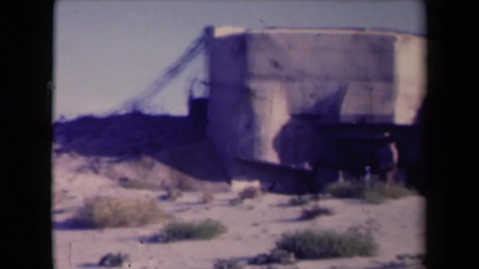 1945 France 8mm frame with a large bunker-like concrete structure and two small figures at its base in harsh sunlight.