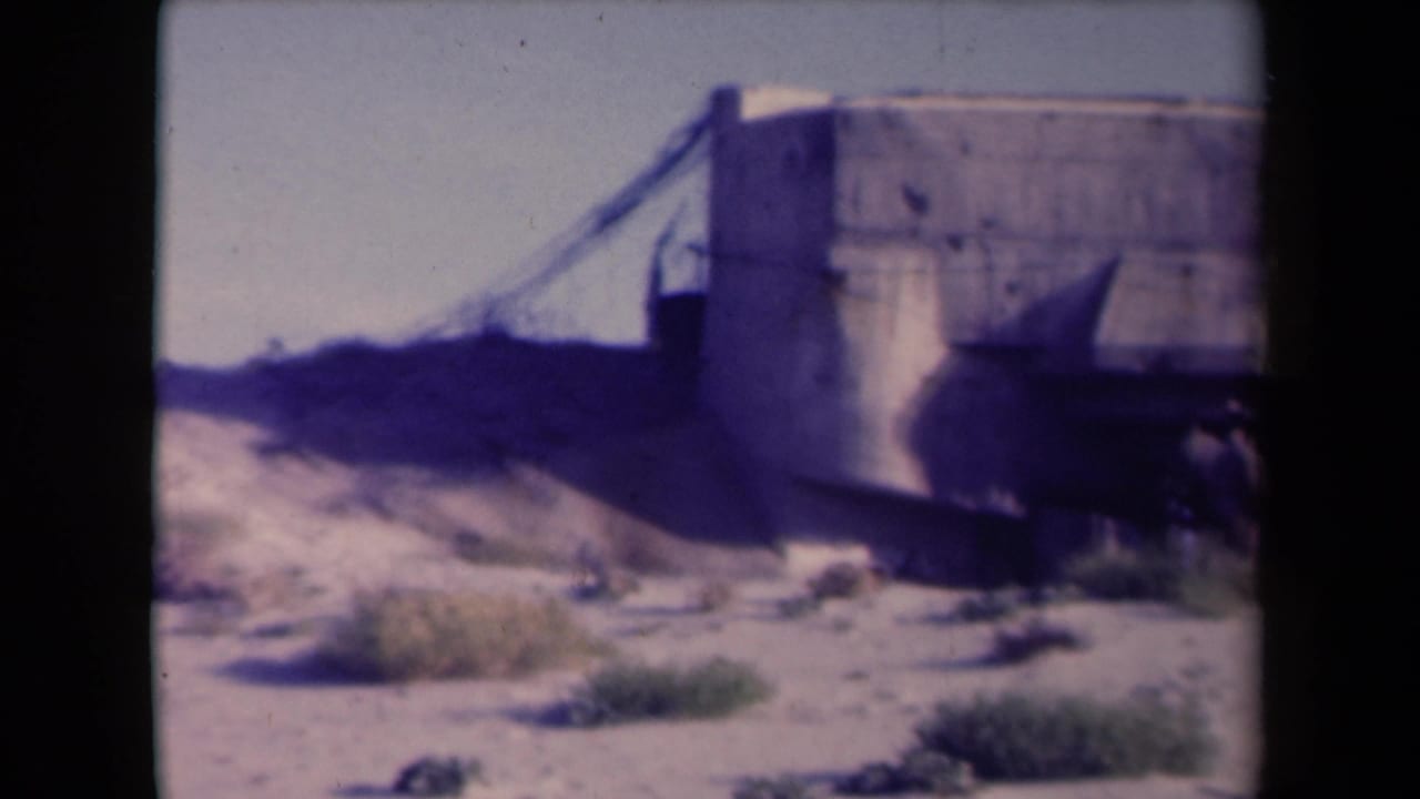 Concrete bunker-like structure in sandy terrain with sparse vegetation and dark film-frame edges.
