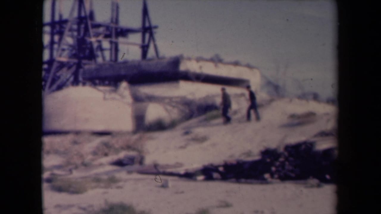 Two men walk near a concrete military structure with metal tower or scaffolding behind it.