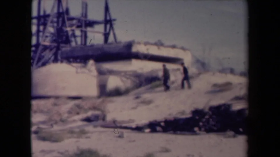 1945 France frame with two distant men crossing sandy ground beside a bunker-like concrete installation and metal tower.