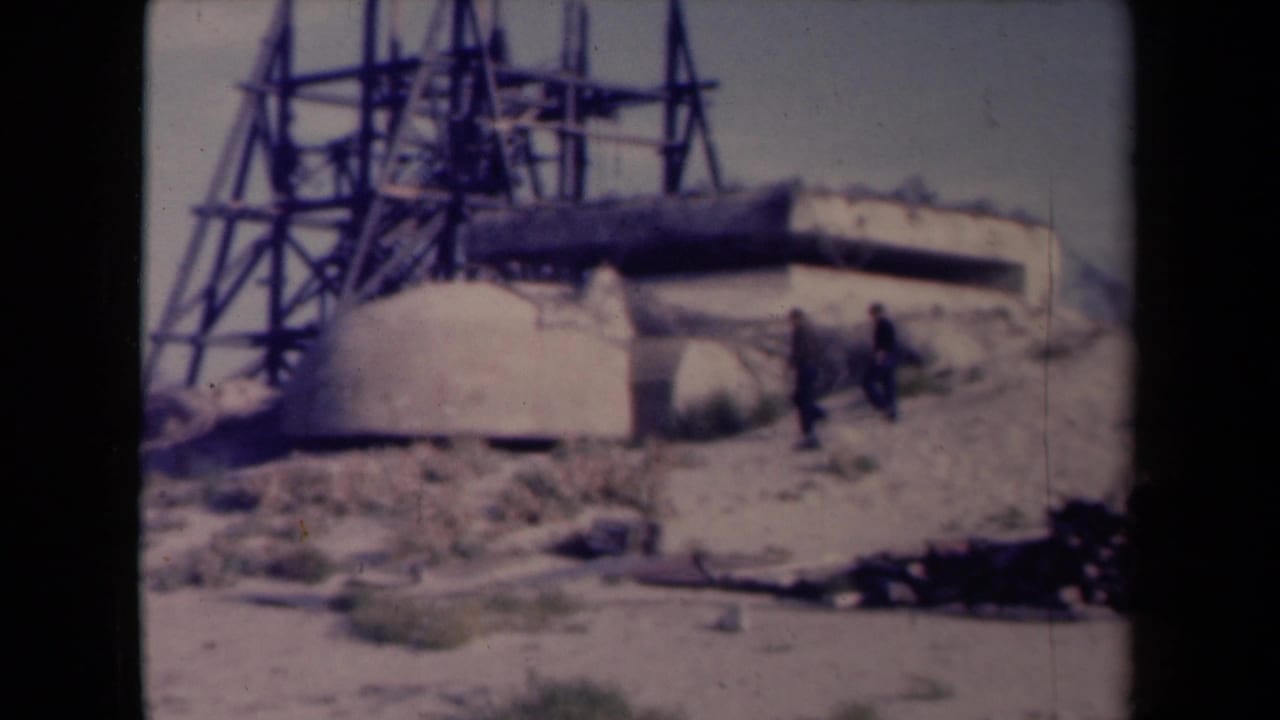 1945 France 8mm frame showing two men near a concrete defensive installation with a tall metal framework rising behind it.