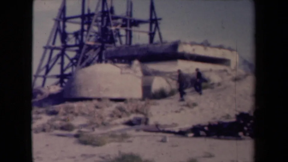 1945 France 8mm frame showing two men near a concrete defensive installation with a tall metal framework rising behind it.