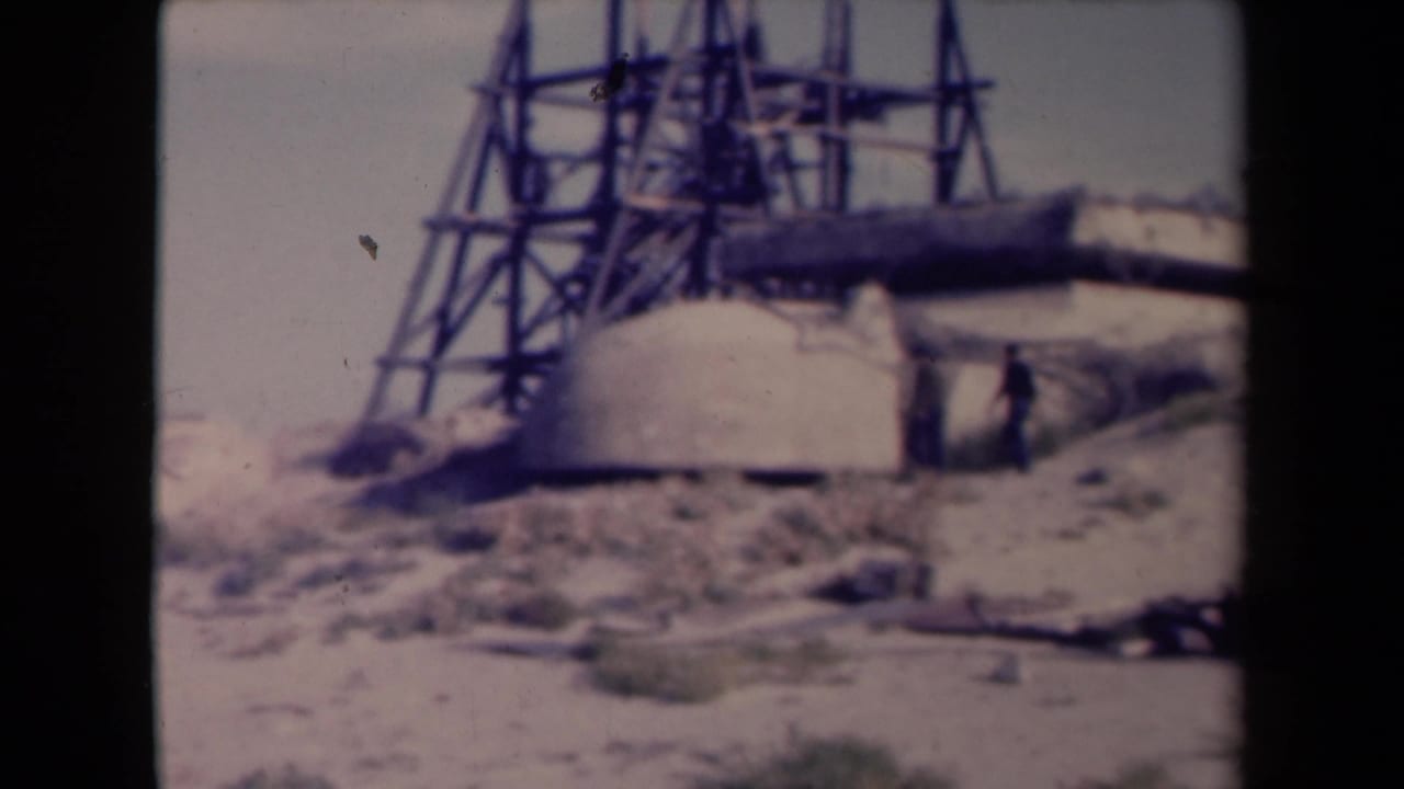 1945 France frame showing two distant men beside a concrete defensive structure, with sandy ground and metal scaffolding visible.