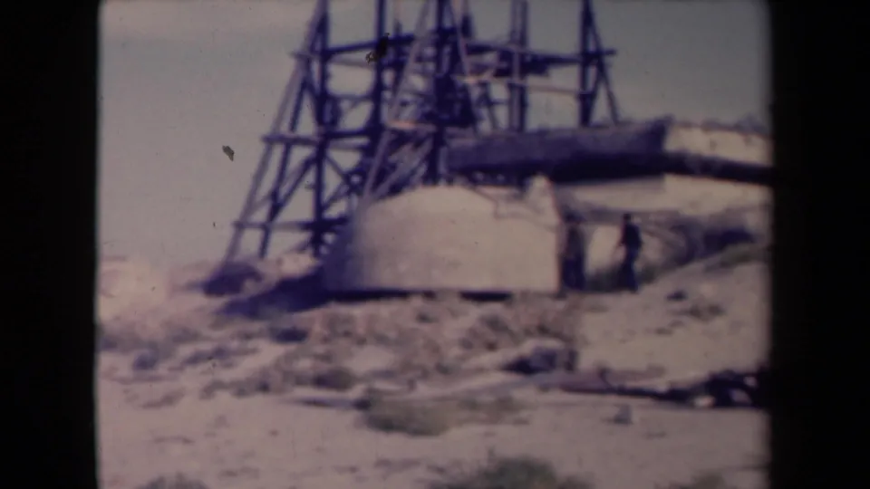 1945 France frame showing two distant men beside a concrete defensive structure, with sandy ground and metal scaffolding visible.
