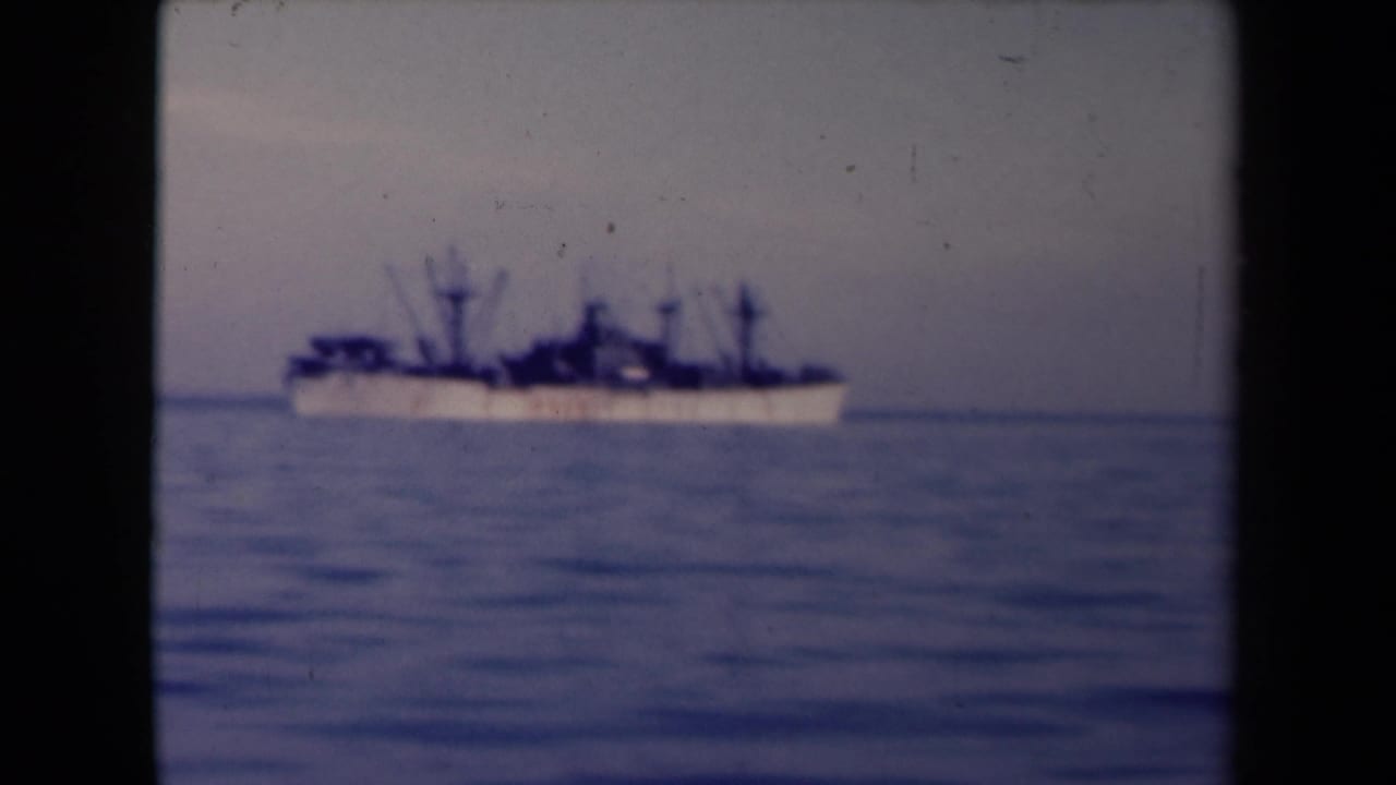 1945 France coastal home-movie frame with a large distant ship offshore on calm water under a muted sky.