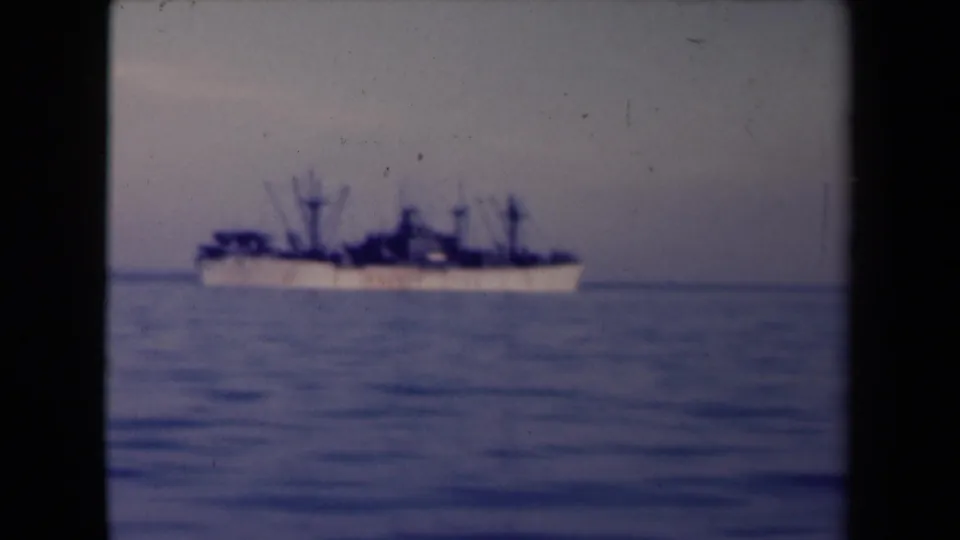 1945 France coastal home-movie frame with a large distant ship offshore on calm water under a muted sky.