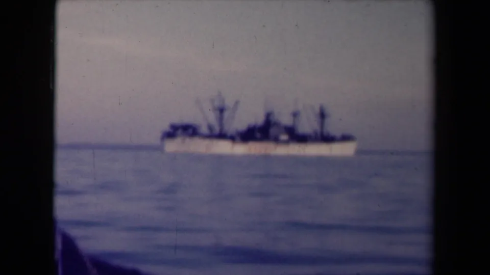 Soft 8mm frame of a large ship at sea in France, with visible grain, faded color, and a dark foreground shape at lower left.