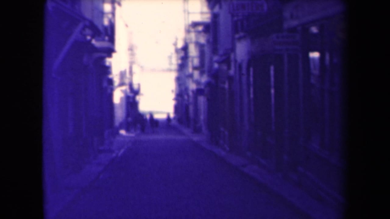 1945 France frame looking along a shaded village street, with buildings on both sides and people far ahead.