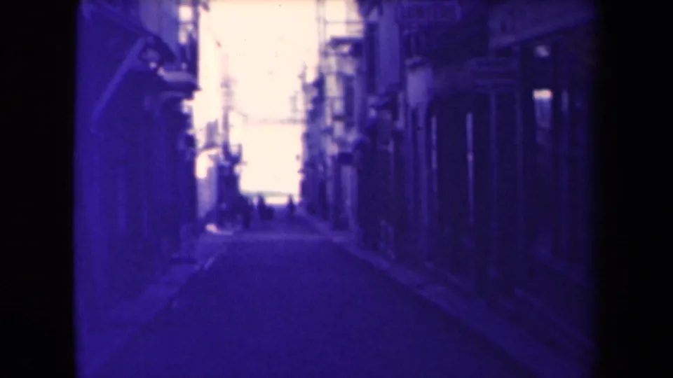 1945 France frame looking along a shaded village street, with buildings on both sides and people far ahead.