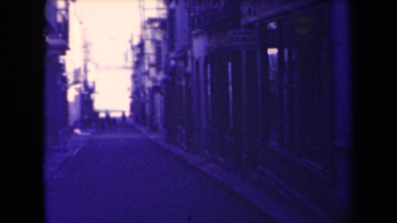 Narrow street with dark buildings, distant figures, bright sky, and visible film edge shadows.