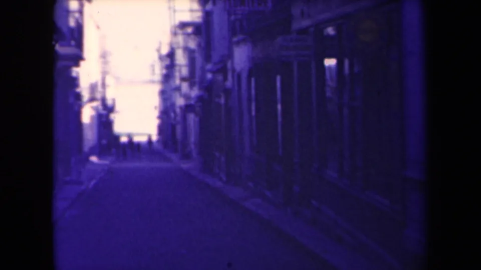 1945 France street view on 8mm film, with a narrow lane, shadowed facades, distant pedestrians, and heavy gate-edge darkness.