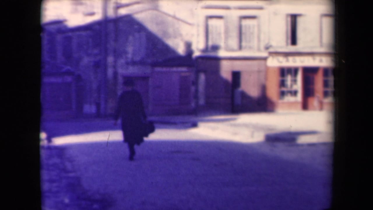1945 France frame showing a dark-clothed pedestrian crossing a sunlit street in front of older buildings and visible shop signage.