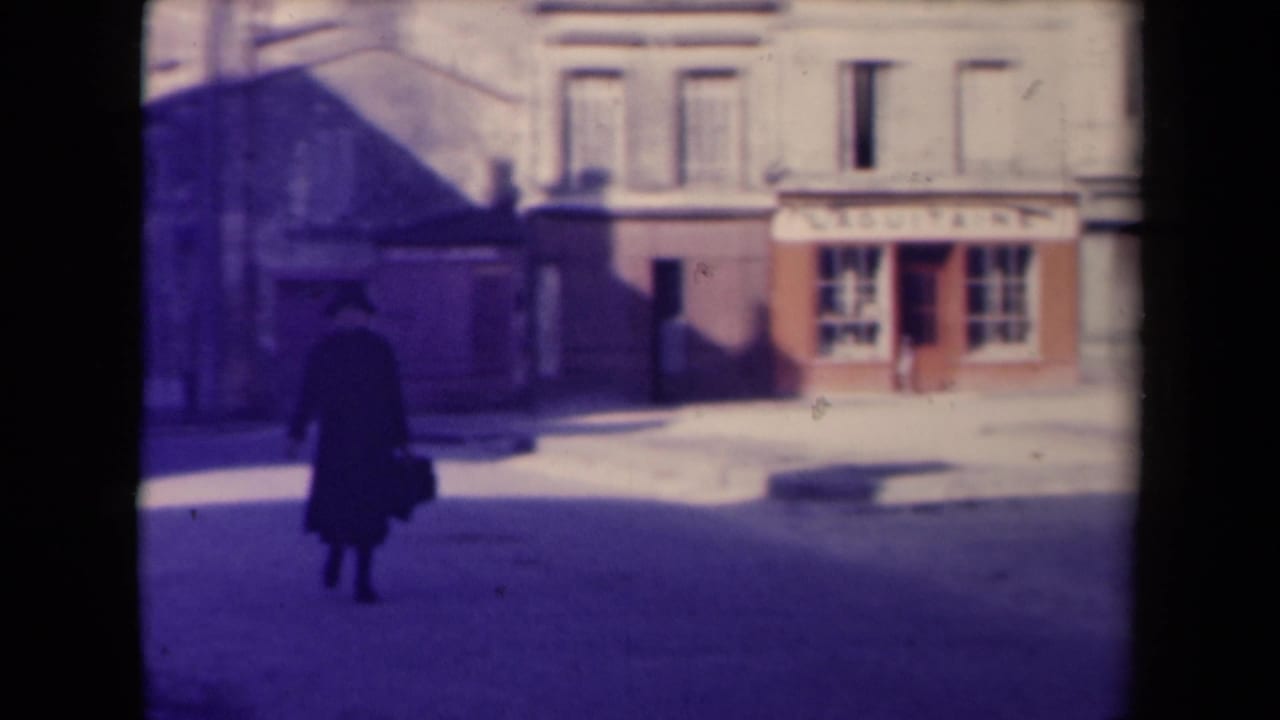 1945 France 8mm frame with a lone dark-clothed figure crossing an open street before pale facades and a shop sign.