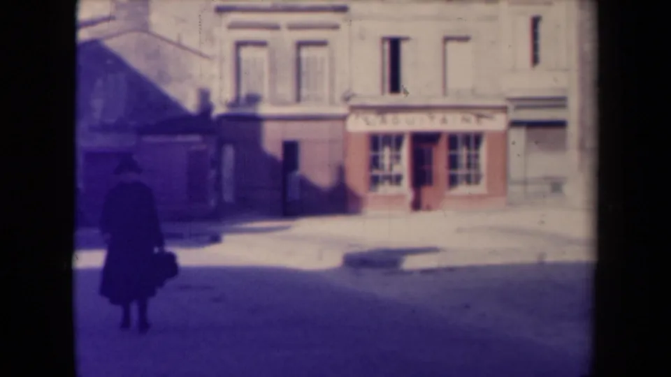 In 1945 France, a man in a dark coat and hat crosses a quiet street beside a shopfront and pale civic-style building.