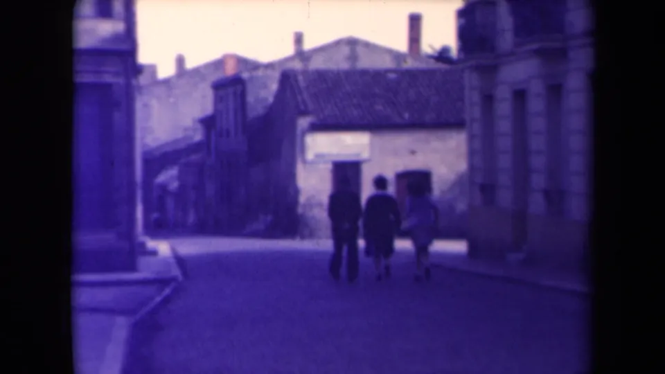 Three pedestrians walk away on a narrow French town street, framed by stone buildings, tiled roofs and soft purple-tinted film color.