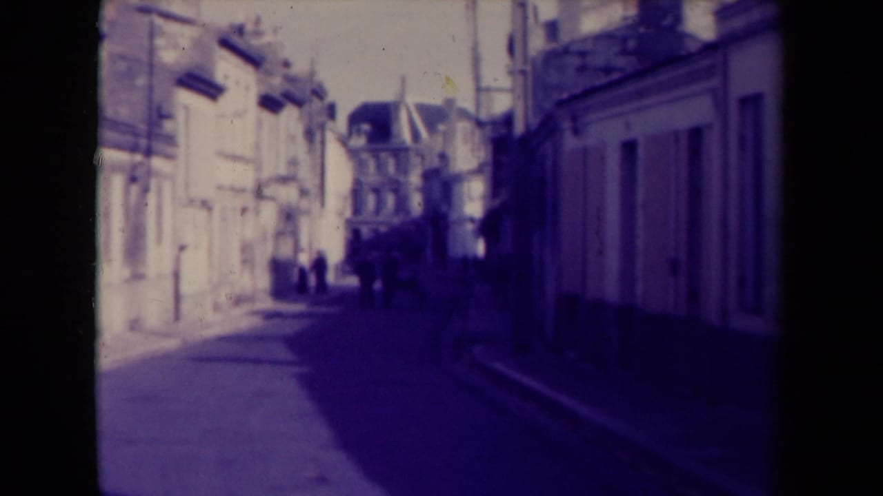 1945 France 8mm frame showing pedestrians ahead on a narrow street, with sunlit building fronts on one side and deep shadow on the other.