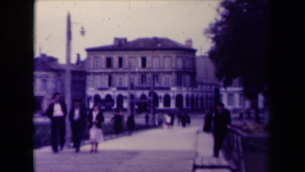 1945 France frame showing pedestrians on a bridge or riverside walkway, railings, a tall column or monument, and a large building beyond.