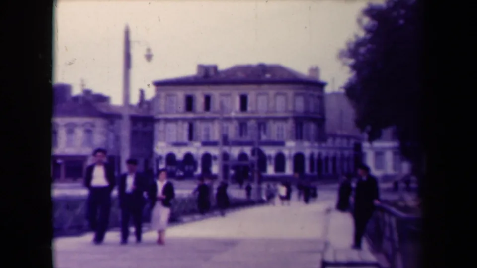 In 1945 France, a small group walks toward the camera on a broad promenade beside historic-looking buildings and a tall monument.