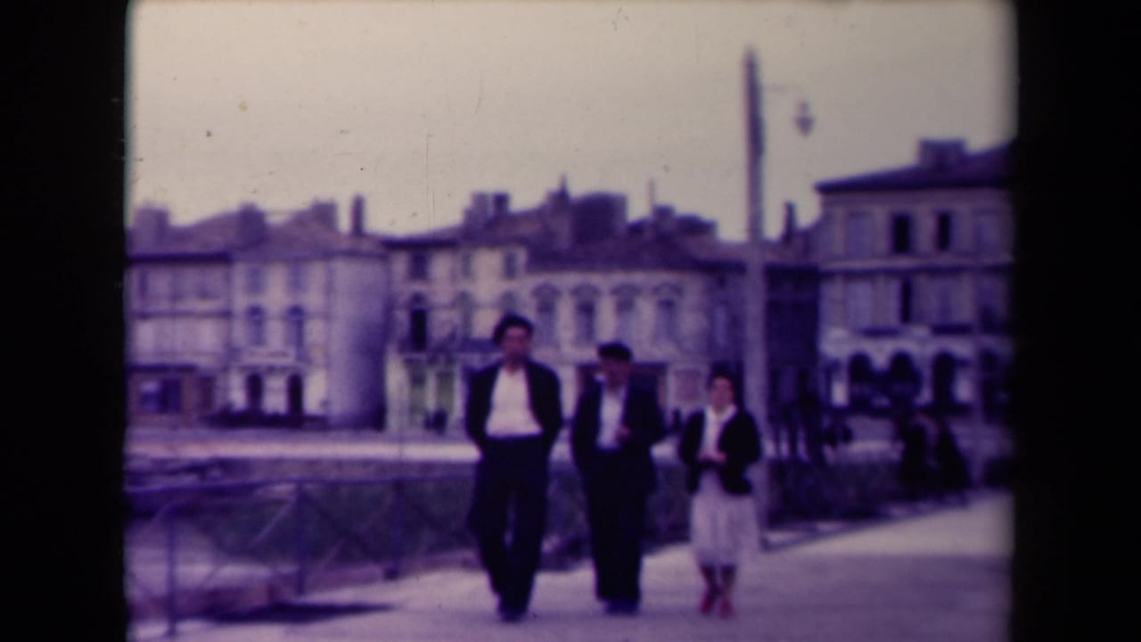 Three people walk along a waterside bridge with houses behind them.
