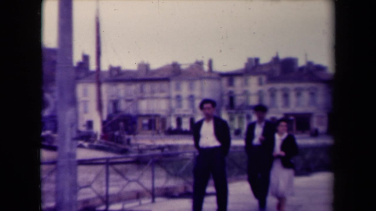 Three pedestrians pass near a tall post or monument on a waterside walkway in France, with houses and railings behind them.