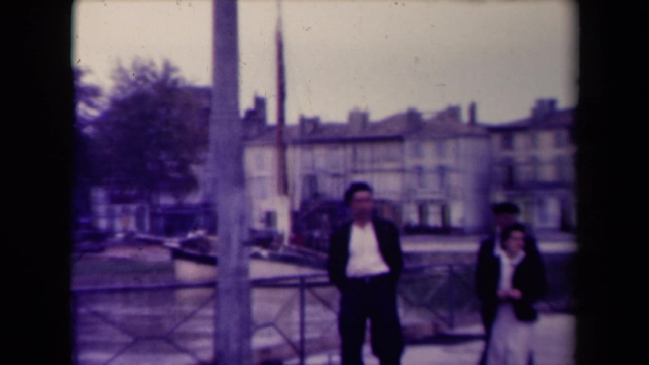 Two walkers are prominent near the right side of the frame as the waterside railing, water, and French town buildings remain visible.