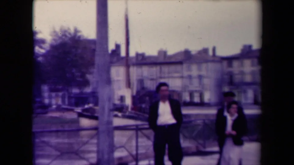 Two walkers are prominent near the right side of the frame as the waterside railing, water, and French town buildings remain visible.