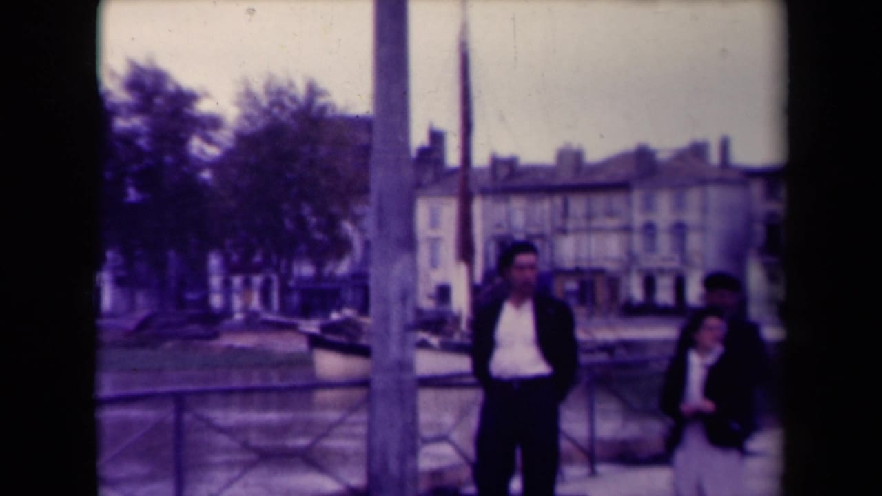 Two people stand near a riverside railing with buildings and trees across the water.