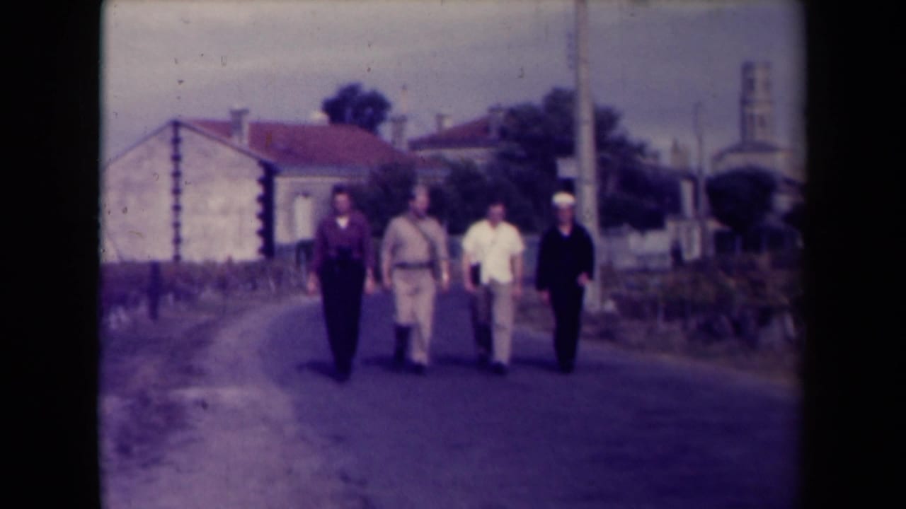 The group of four men continues walking toward the camera in a rural French setting, framed by houses, roadside vegetation, and a distant tower.