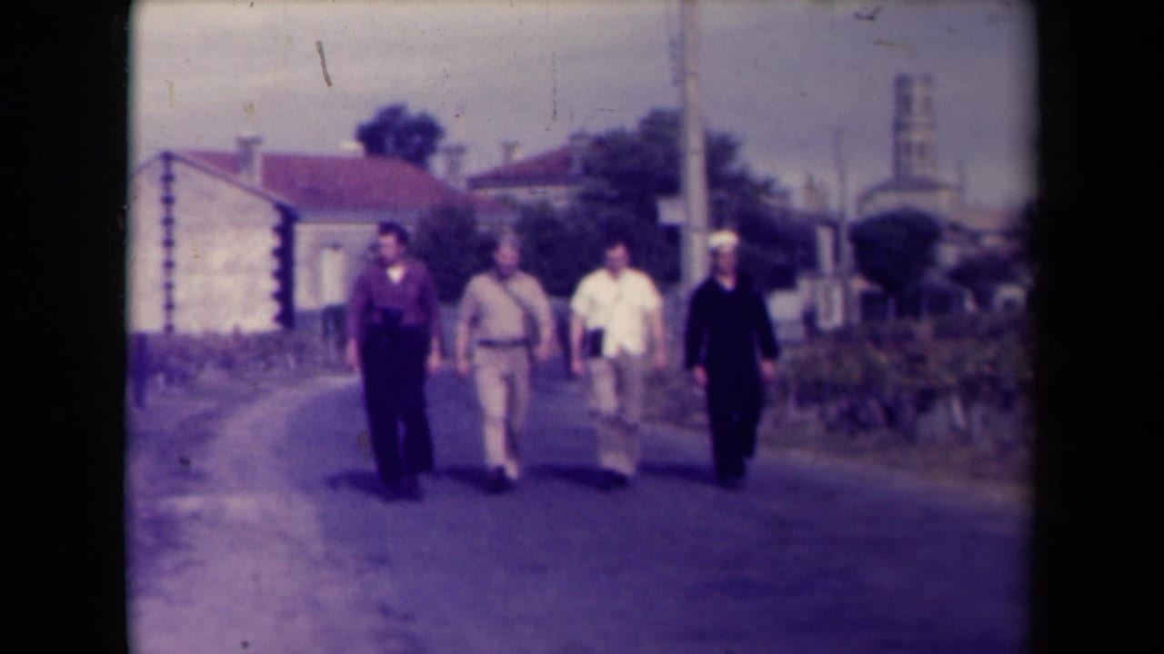Four men in civilian or uniform-like clothing walk together on a village road in France, with buildings, a utility pole, and a distant tower visible.
