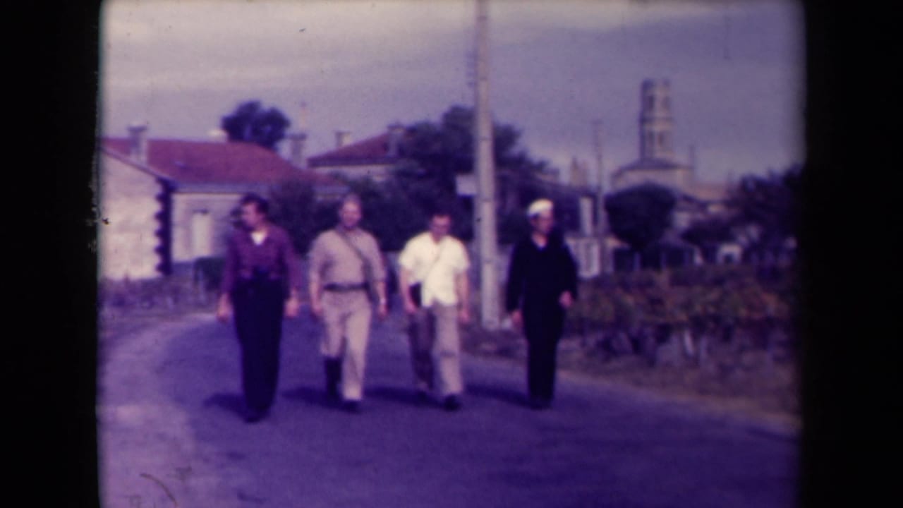 Four men walk toward the camera on a rural road beside vineyards and low buildings.