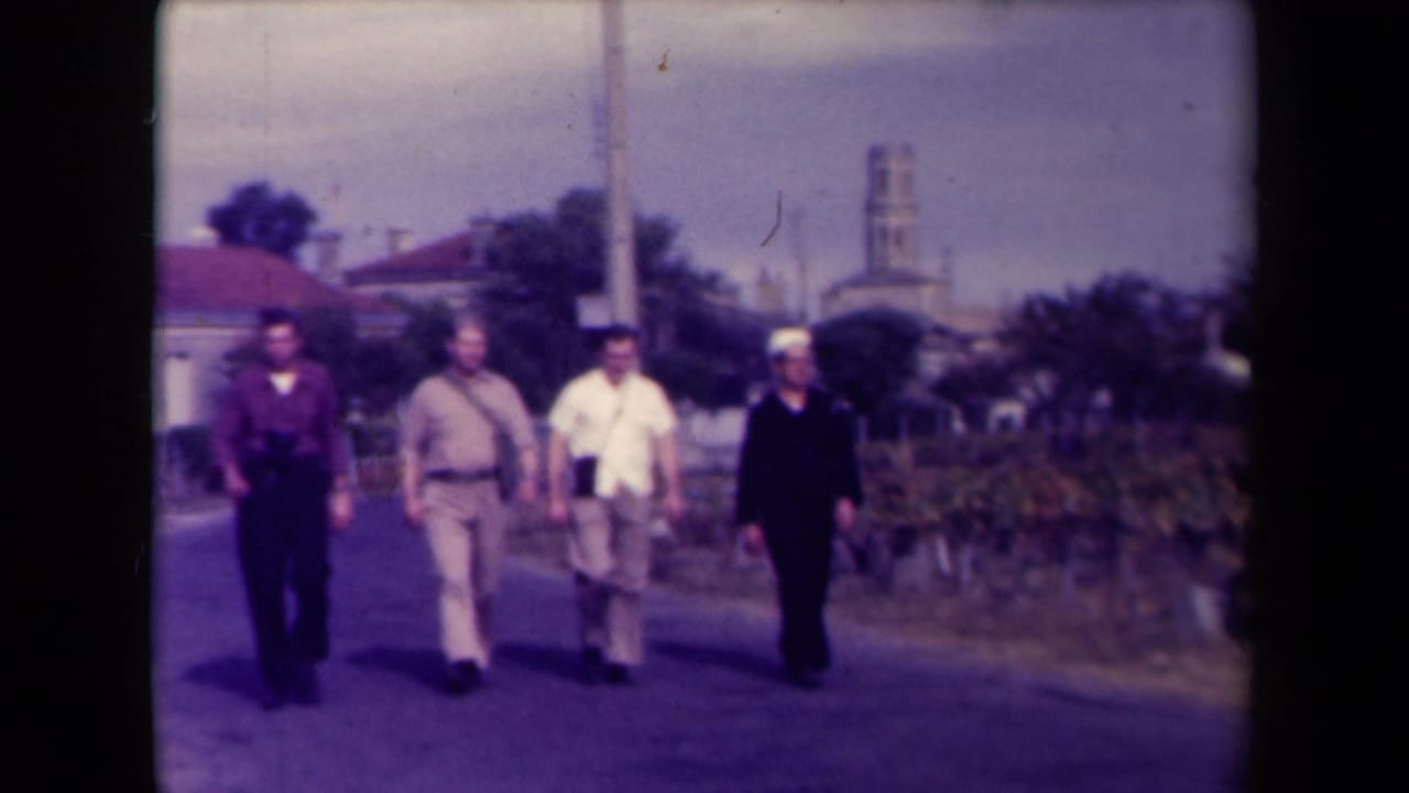 Four men continue walking along the road, with vineyards and a steeple in the background.